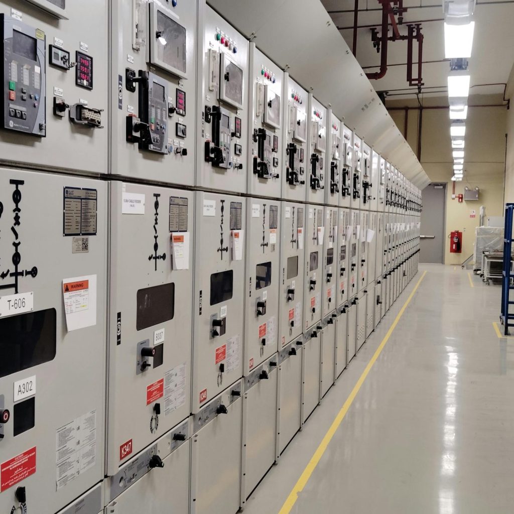 Long row of control panels in an industrial electrical control room, well-lit and spacious.